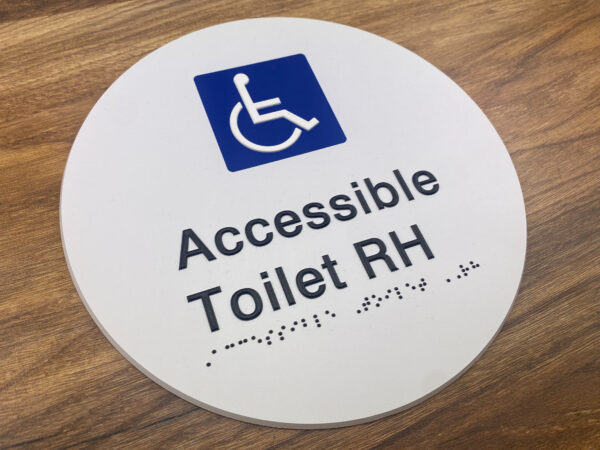 Photo of an Accessible Toilet RH Braille Sign. White disk with Blue & White international symbol for Access. Black text reads Accessible Toilet RH