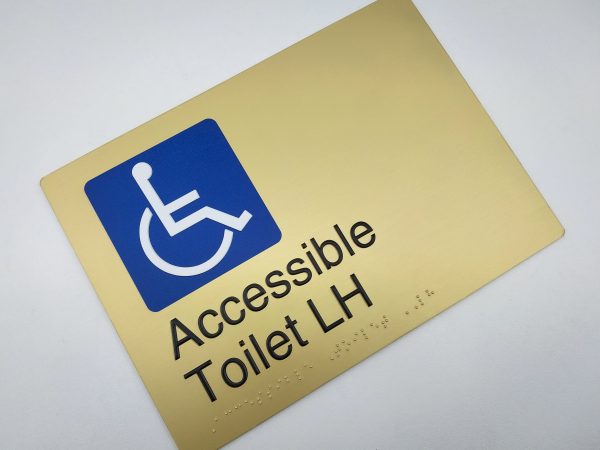 Accessible Toilet LH Braille Sign in Landscape Satin Brass. Brass Sign panel with Blue & White Accessible Symbol & Text that reads Accessible Toilet LH. Braille Text is shown