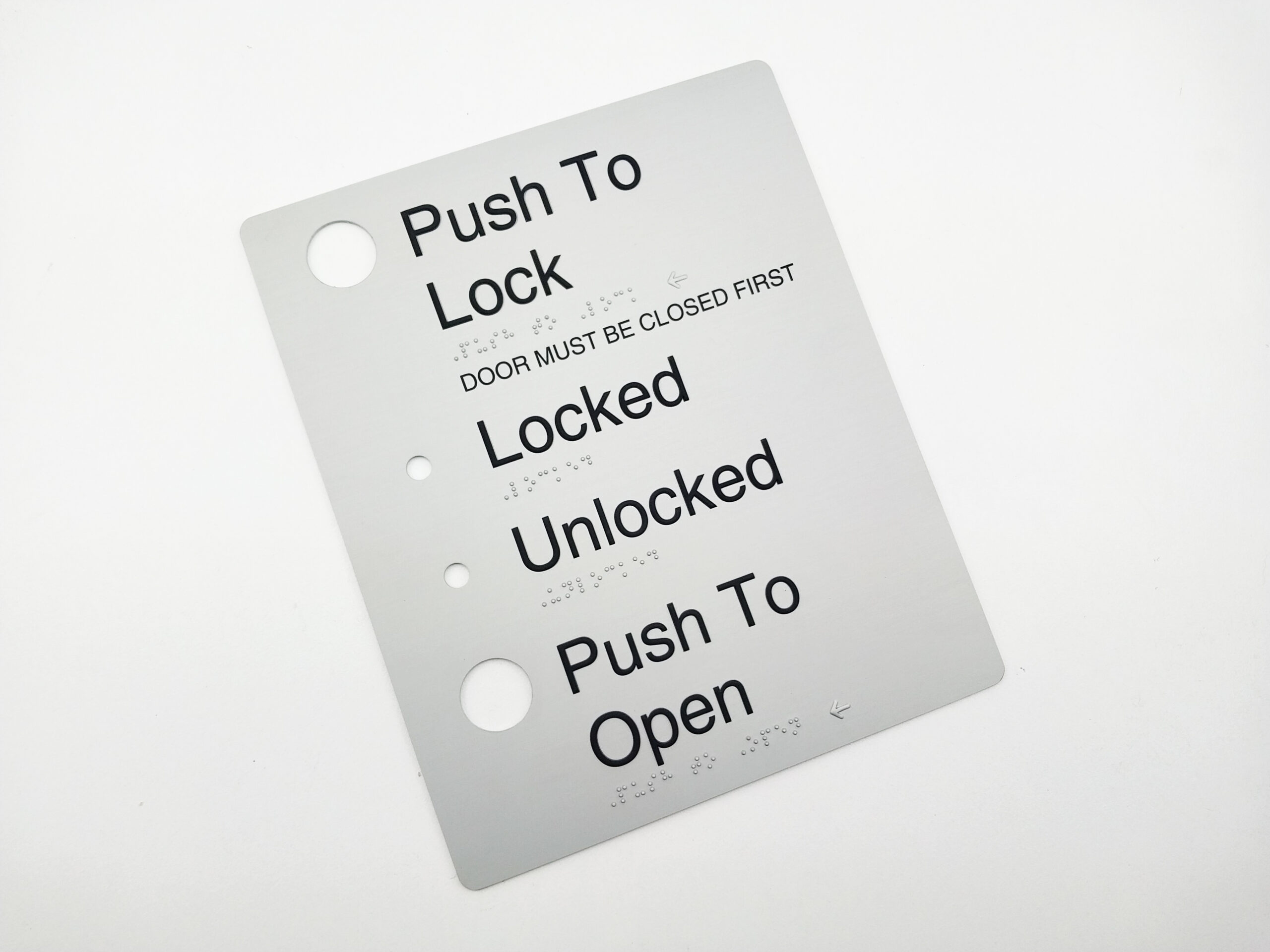 Push to Lock, Automatic Door Braille Sign in Anodised Aluminium. Silver sign panel with Black Raised text & Button holes. Braille is also shown